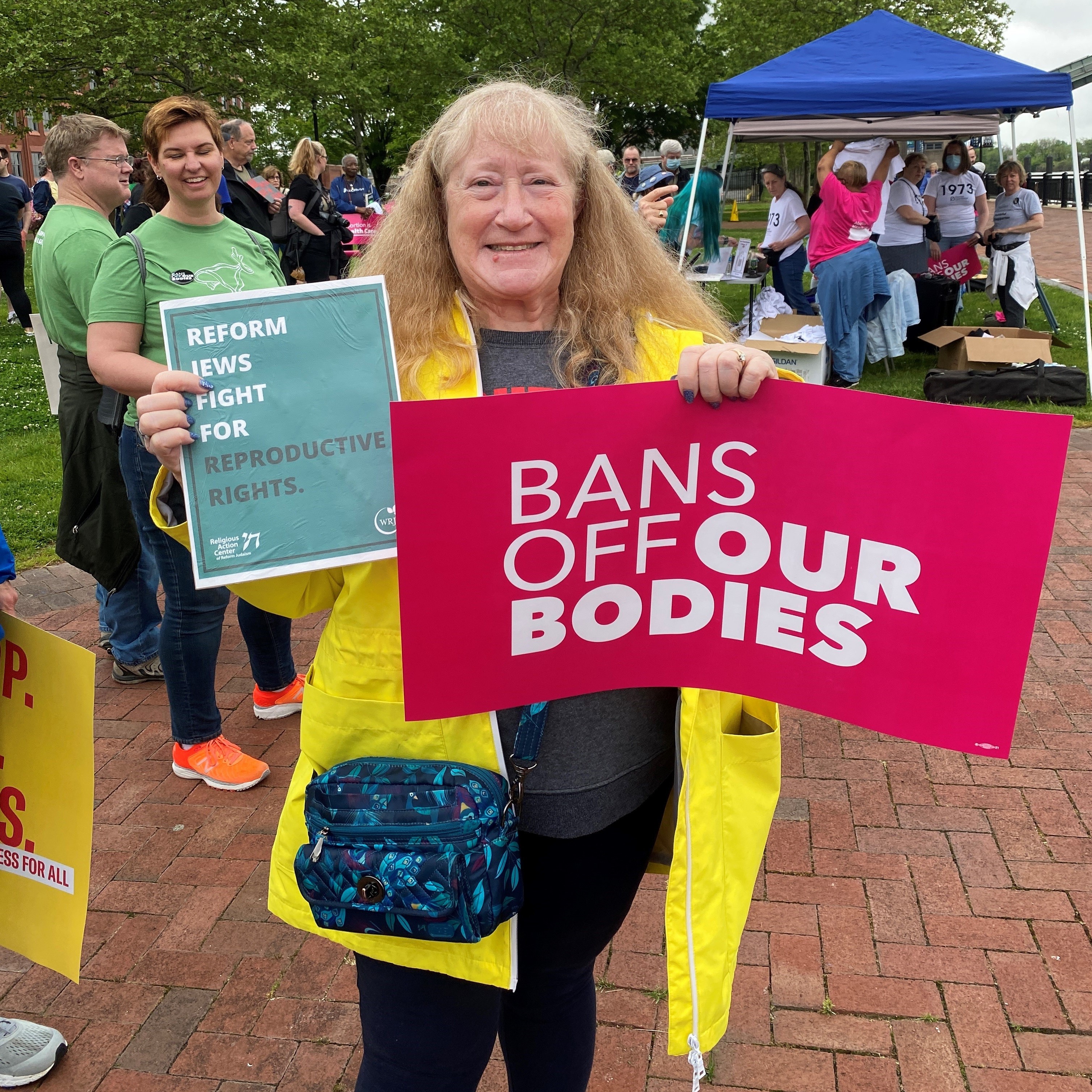 Woman in yellow jacket holding rally signs for "Bans off our Bodies" and Reform Jews for Reproductive RIghts