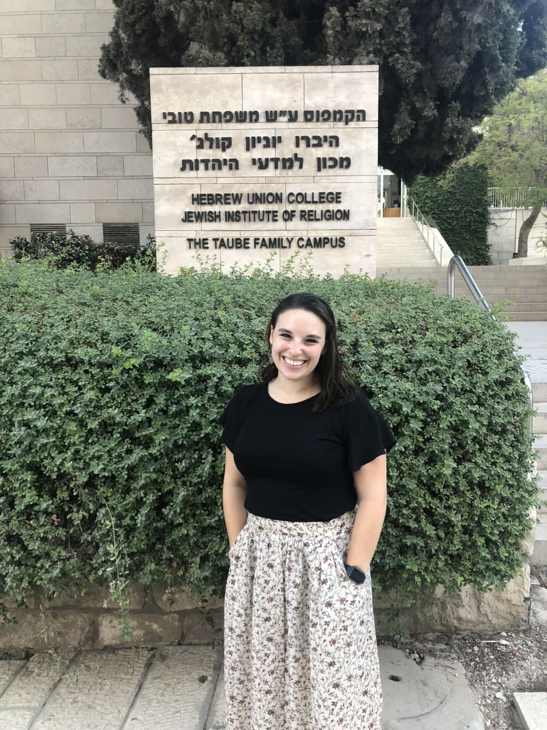 A photo of a woman in a black shirt and printed skirt in front of the HUC sign