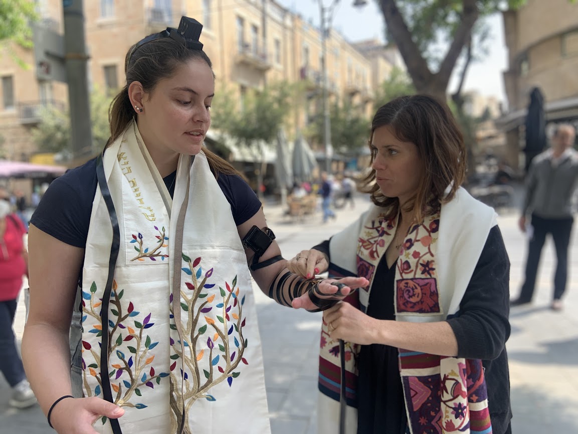One woman with short brown hair putting on tefillin for another woman. 