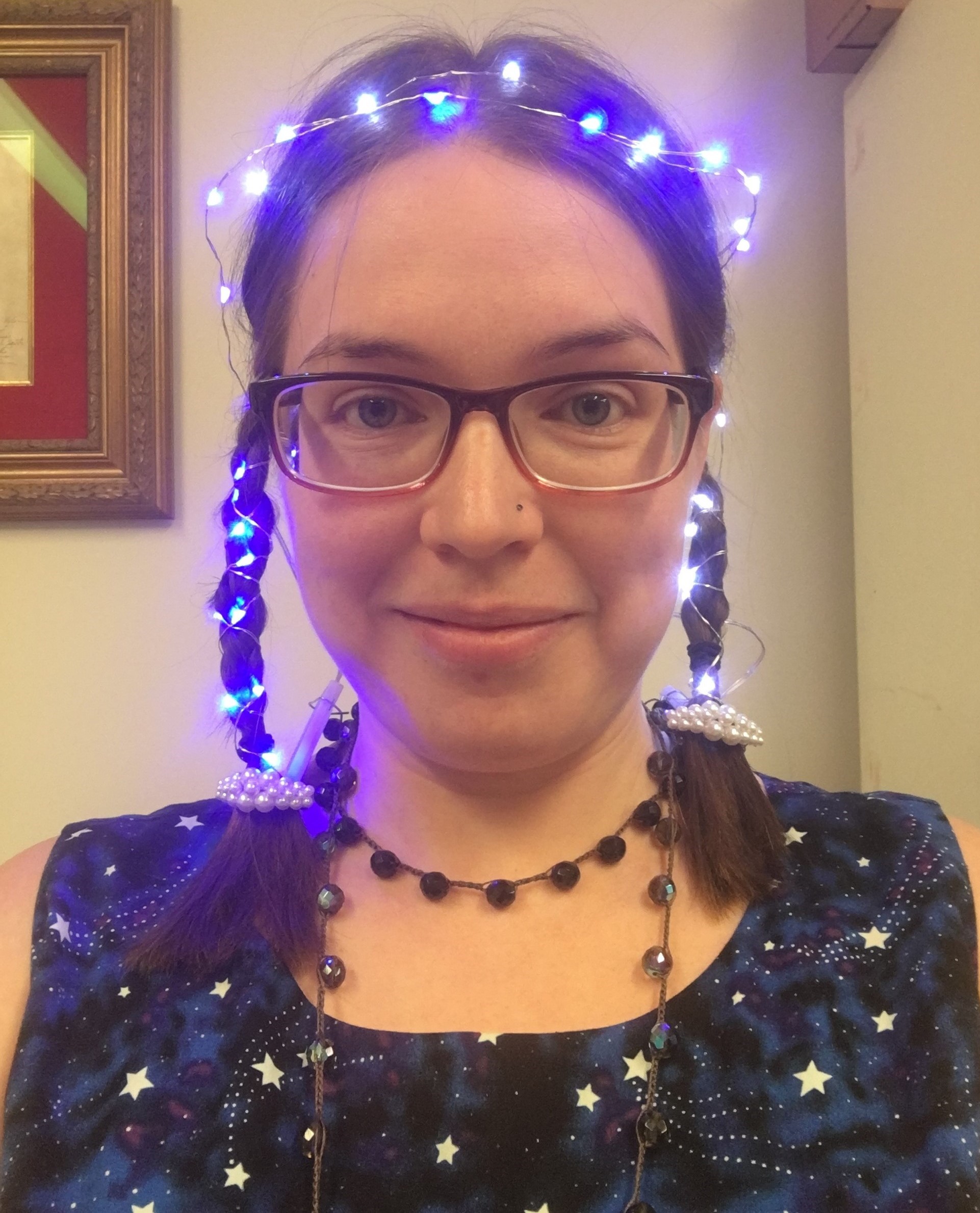 A woman with wearing glasses and brown, shoulder-length hair with two braids and fairy lights woven around her head and braids. She is wearing a dark blue shirt with pale yellow stars.  