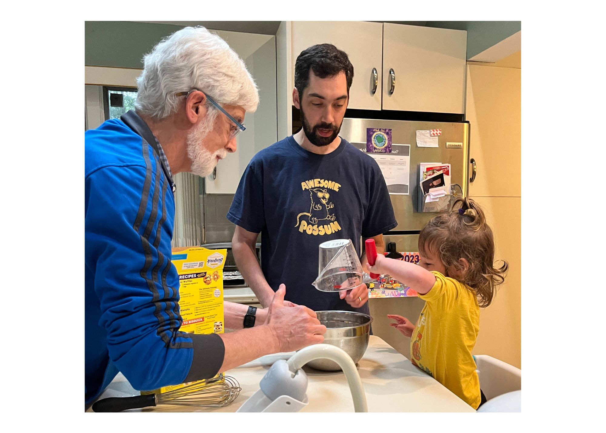A man with white hair and blue shirt, a man with brown hair and beard wearing a black t-shirt, and a young girl with light brown hair wearing a yellow shirt work to stir pancake batter in a kitchen. 
