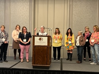 A group of nine women stand on a stage with a podium. 