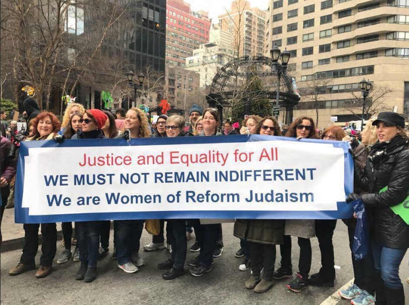 A group of WRJ women in a city holding a white banner with blue border that reads, "Justice and Equality for All, WE MUST NOT REMAIN INDIFFERENT, We are Women of Reform Judaism"