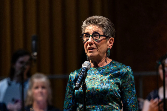 Barbara Ostfeld, a woman with short gray hair, round black glasses, and a peacock-printed shirt, stands in front of a microphone