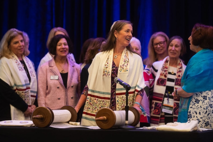 Karen Goldberg wearing a cream multicolored tallit standing in front of an open Torah