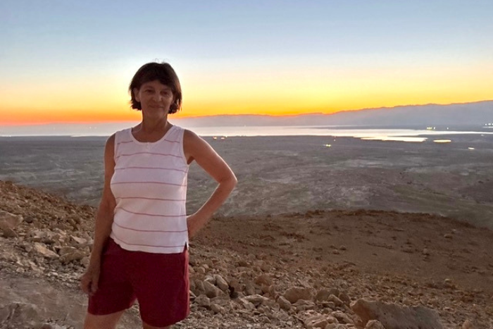 woman with white tank top and maroon shorts standing on mountain with Dead Sea in the background