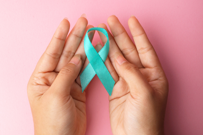 hands holding a teal ribbon in front of a pink background