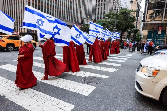 A line of women in red robes and white hats from the Handmaids Tale. They are carrying Israeli flags.
