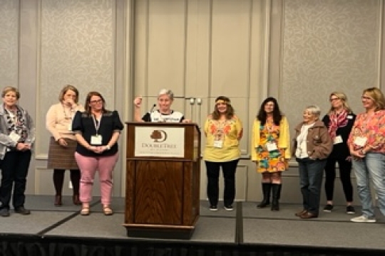 A group of nine women stand on a stage with a podium.