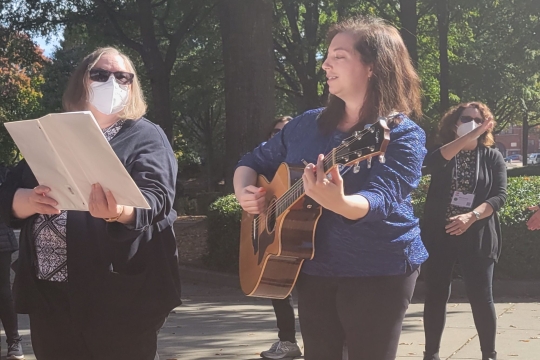 Rabbi Marla J Feldman in sunglasses and a COVID-19 mask hold a white rectangular lyrical book while Kyra Goldman looks on and plays guitar. 