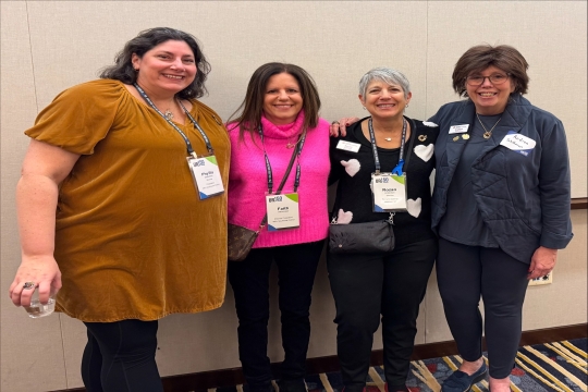 A group of four women standing together against a beige wall. 