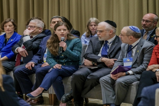 Rabbi Liz P.G. Hirsch speaking into a microphone surrounded by mostly men