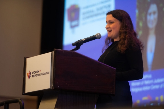 girl with brown hair wearing black long sleeve dress speaking at podium into microphone