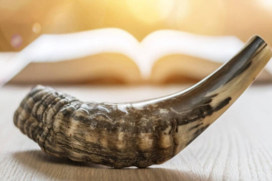 Closeup of a shofar with an open prayerbook in the back of the shot